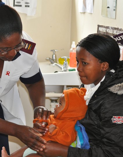 Woman sitting in a chair with an infant in her lap while a health worker examines the infant.