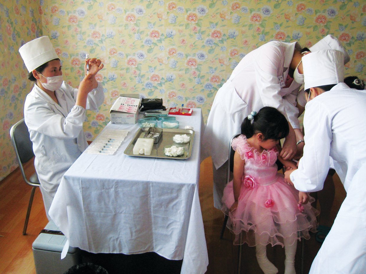 Two medical workers hold a little girl's arm while another draws vaccine into a syringe from a vial at a table covered with vaccination supplies.