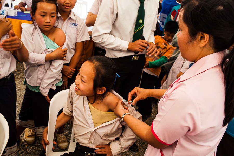 A schoolgirl sits in a white plastic chair as a health worker administers vaccine in the girl's arm.