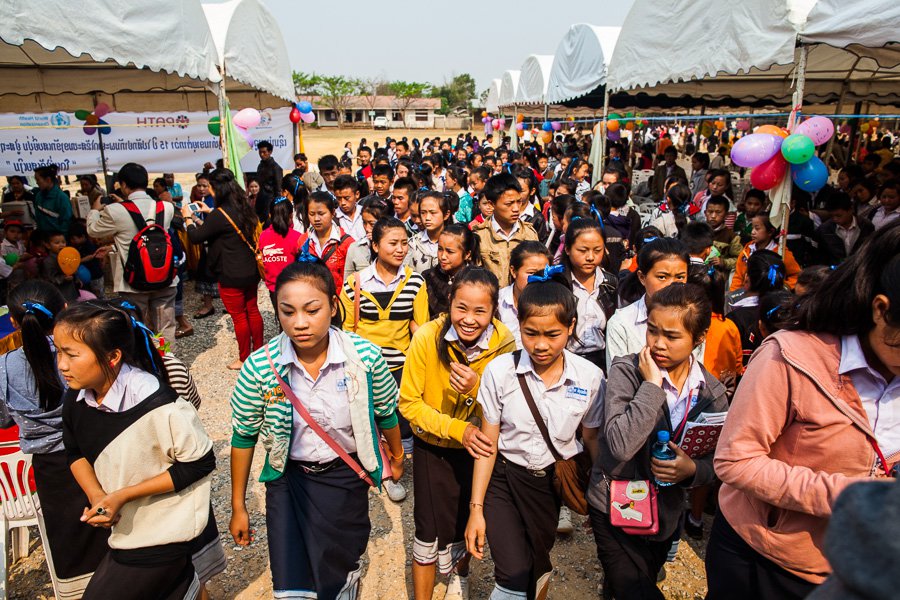 A large crowd of schoolchildren walks through white awnings. A banner behind them advertises a vaccination campaign.