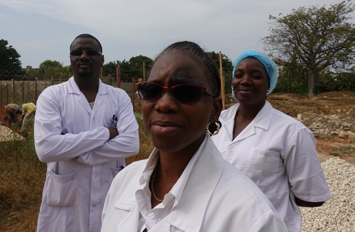 Three people dressed in lab coats standing in a field with workers in the background.
