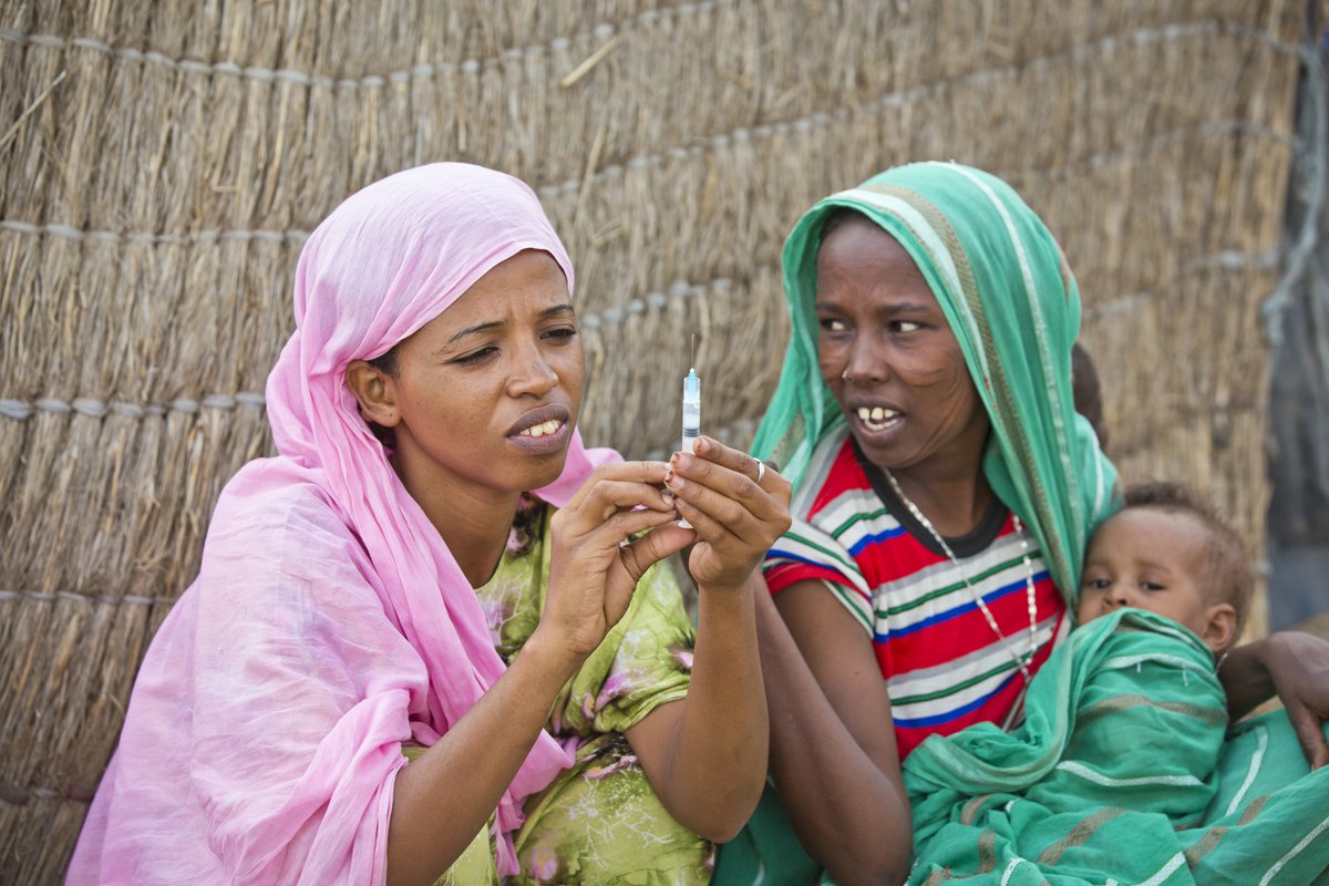A woman in pink head scarf readies a syringe while a woman in green head scarf holding a young child watches.