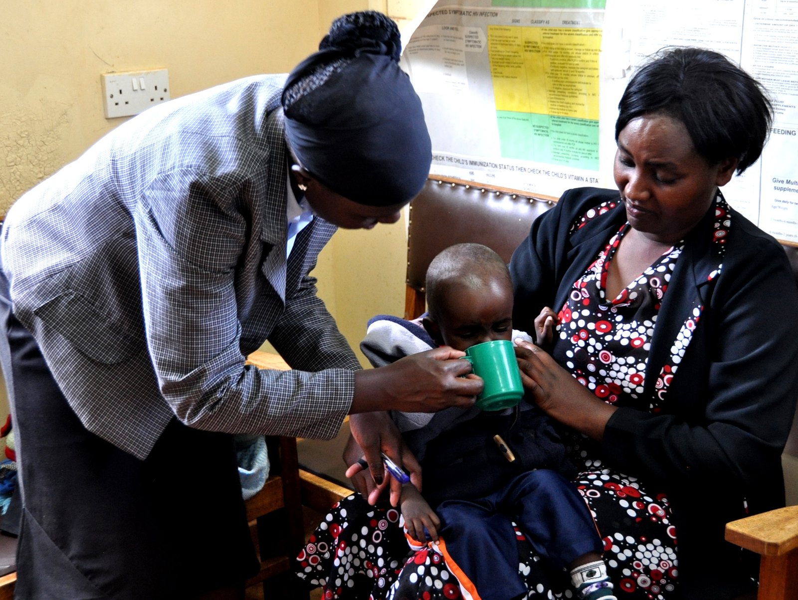 Two women giving a little boy rehydration fluids from a green cup.