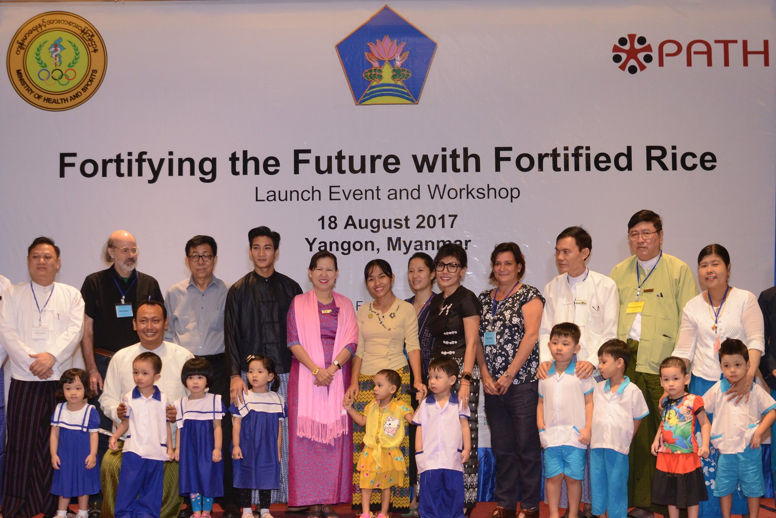 A group of adults and children stand in front of a sign that says "Fortifying the Future with Fortified Rice."
