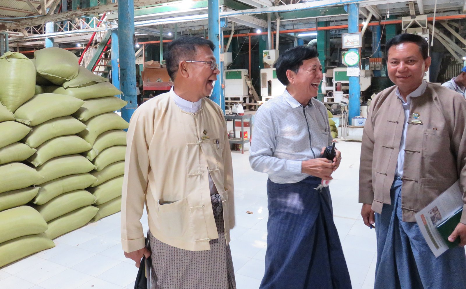 Three men in a warehouse with green bags stacked in the background.