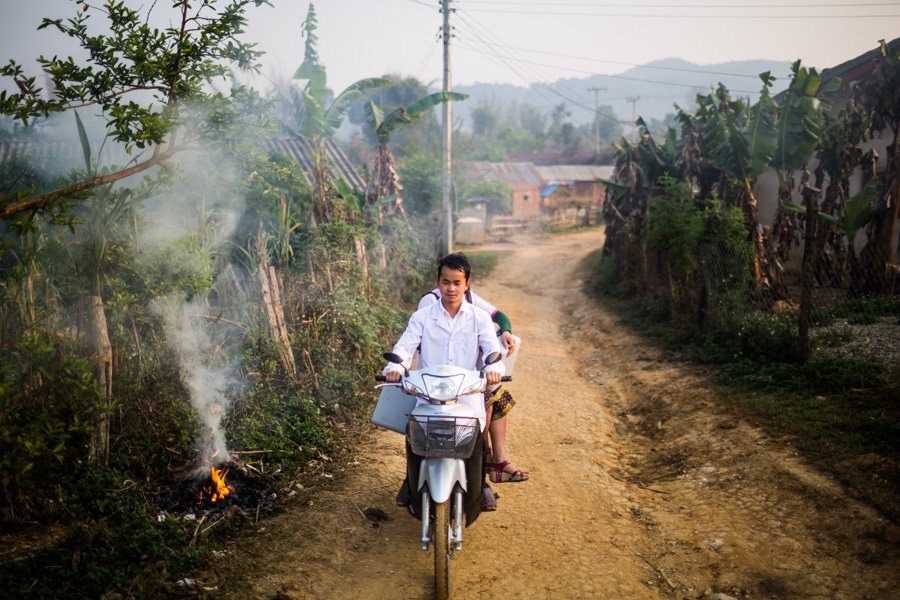 Man drives motorbike on a dirt road. A woman on the back of the motorbike holds a cold storage box.