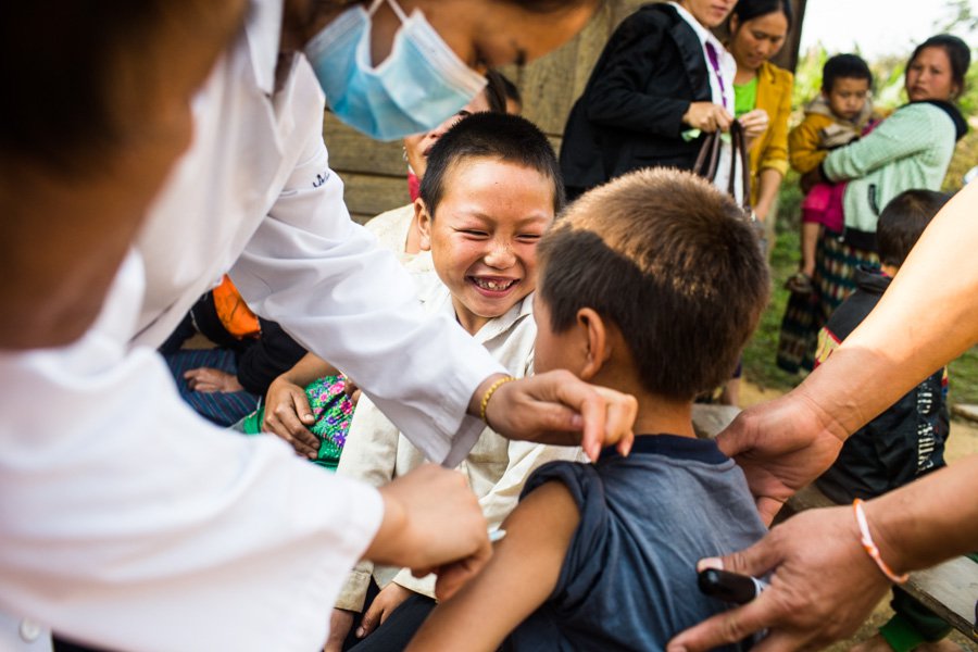 Health worker injects vaccine into the upper arm of a young boy while another smiling boy watches.